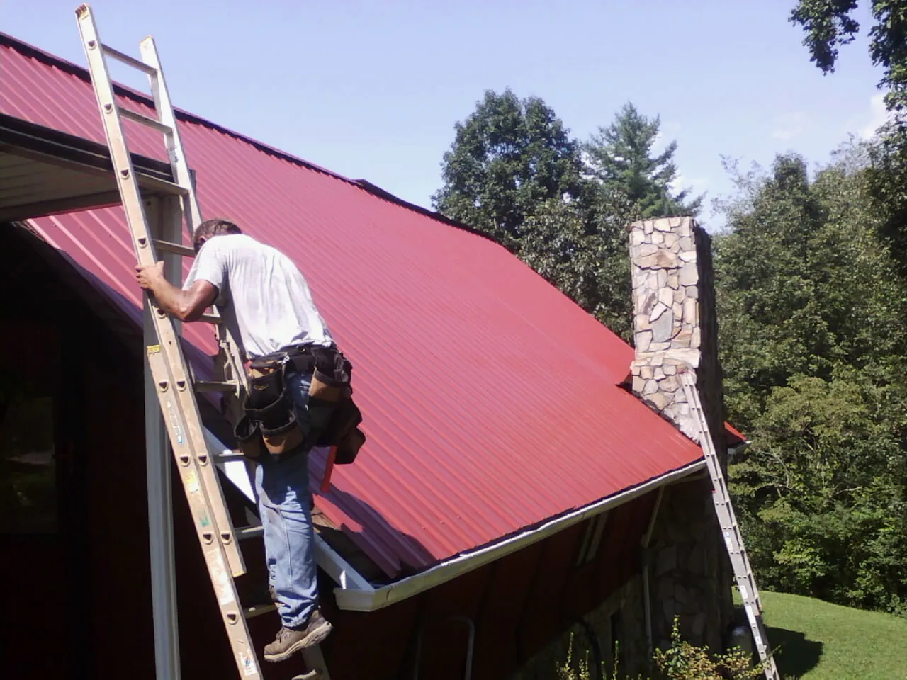 Roofer on a ladder beside a red metal roof and stone chimney — Soffit & Fascia Repair by Roofing Brothers in Martindale